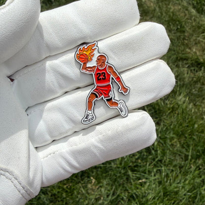 A small golf ball marker shaped like a basketball player in a red jersey with the number 23, holding a flaming basketball, displayed on a white leather glove against a green grass background.