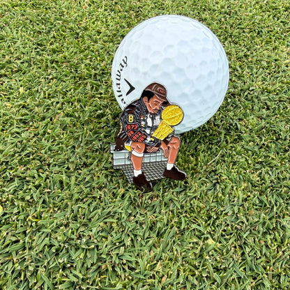 A enamel Mamba Pin Creatures golf ball marker holding a gold trophy sits in front of a white golf ball on a green grass background.