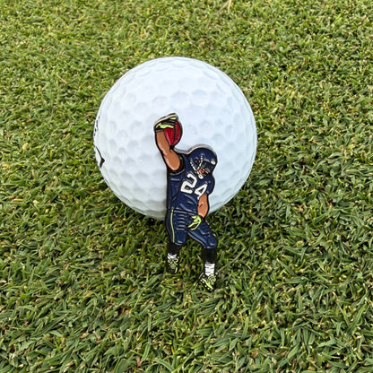 A small enamel golf ball marker shaped like a football player in a navy uniform with the number 24, standing in front of a white golf ball on green grass.